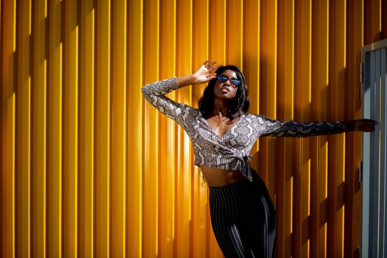 Stylish black woman in crop top and sunglasses posing outdoors against a yellow wall.